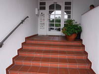 Man sieht die Treppe zum Haupteingang, farblich ( rot ) abgesetzt. Links an der Treppe befindet sich ein Handlauf, der aber frühzeitig endet